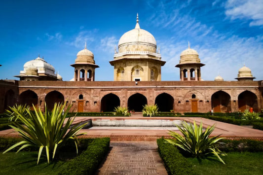 A historic Indian palace with Mughal-style domes and arched corridors, surrounded by manicured gardens and a central fountain under a clear blue sky.