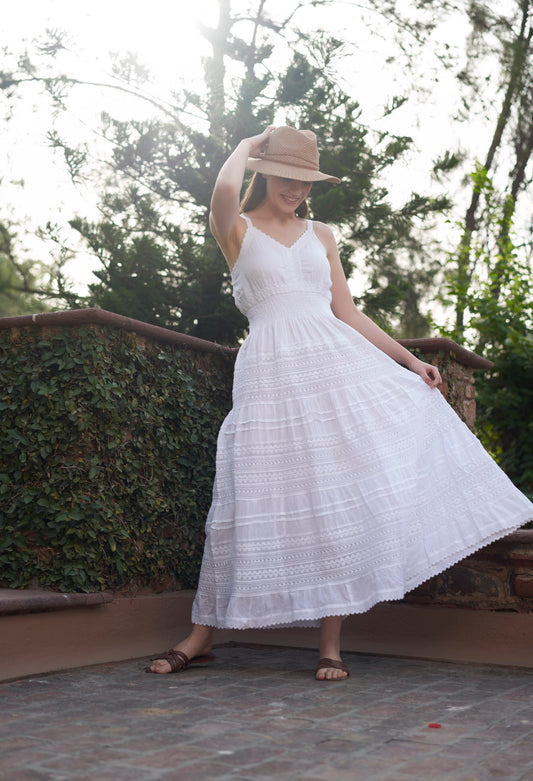 Woman in a white dress and hat standing outdoors with greenery in the background