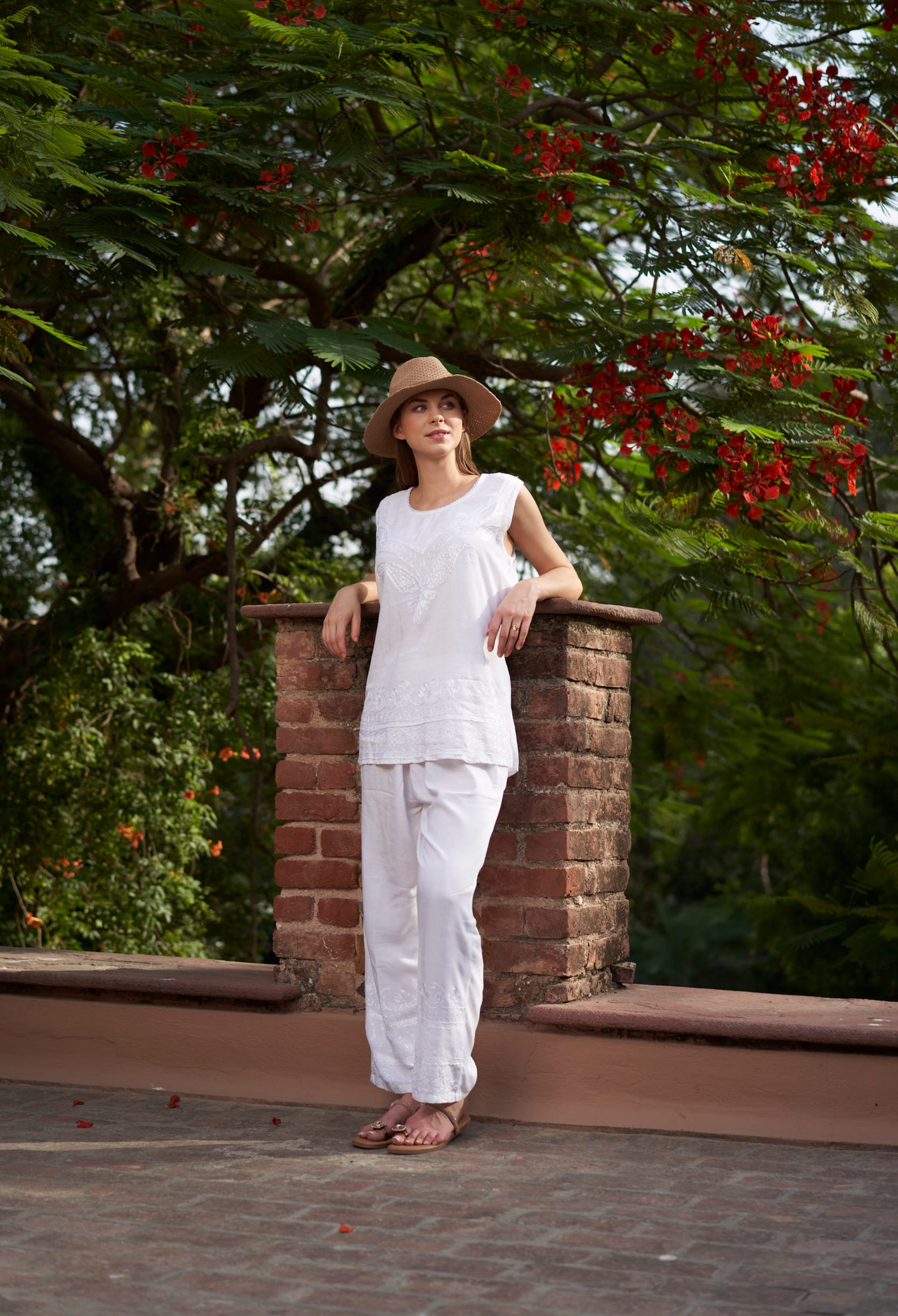 Woman in white outfit and hat leaning against a brick wall with greenery in the background