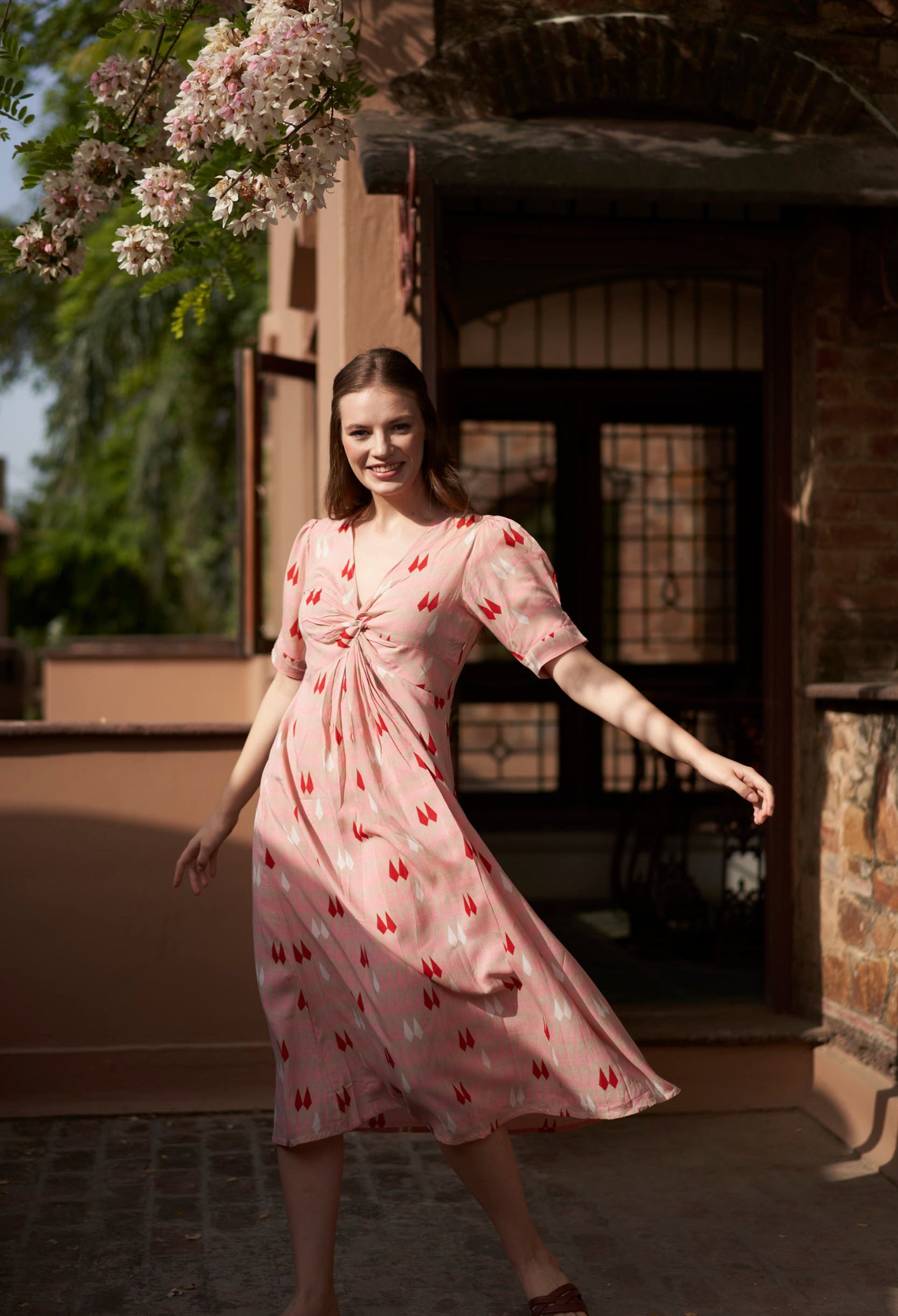 Woman in a pink dress with red patterns standing in front of a building with flowers.