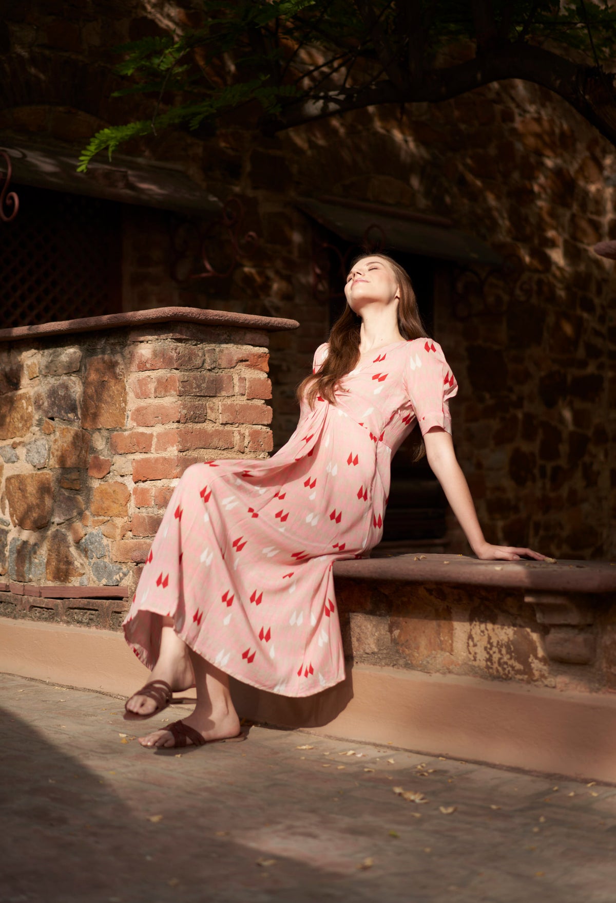 Woman in a pink dress with red patterns sitting on a stone ledge outdoors.