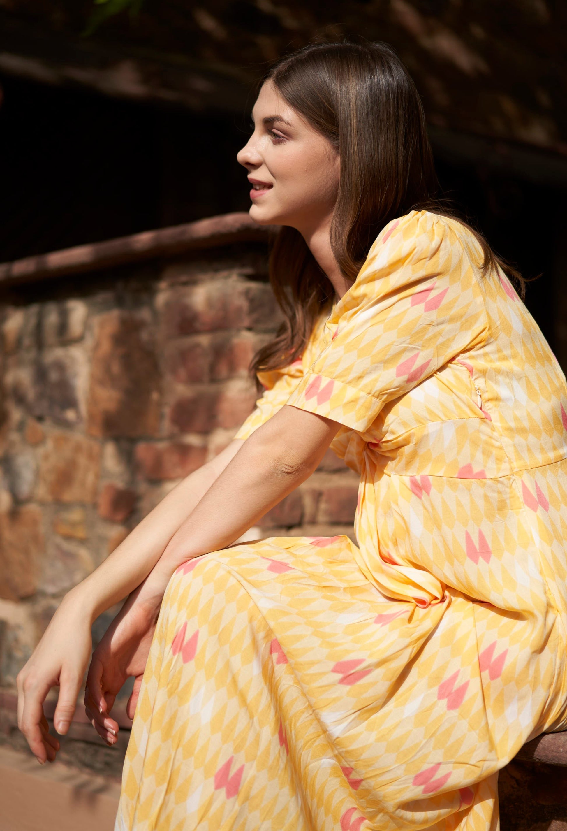 Woman in a yellow dress with pink patterns sitting against a stone wall.