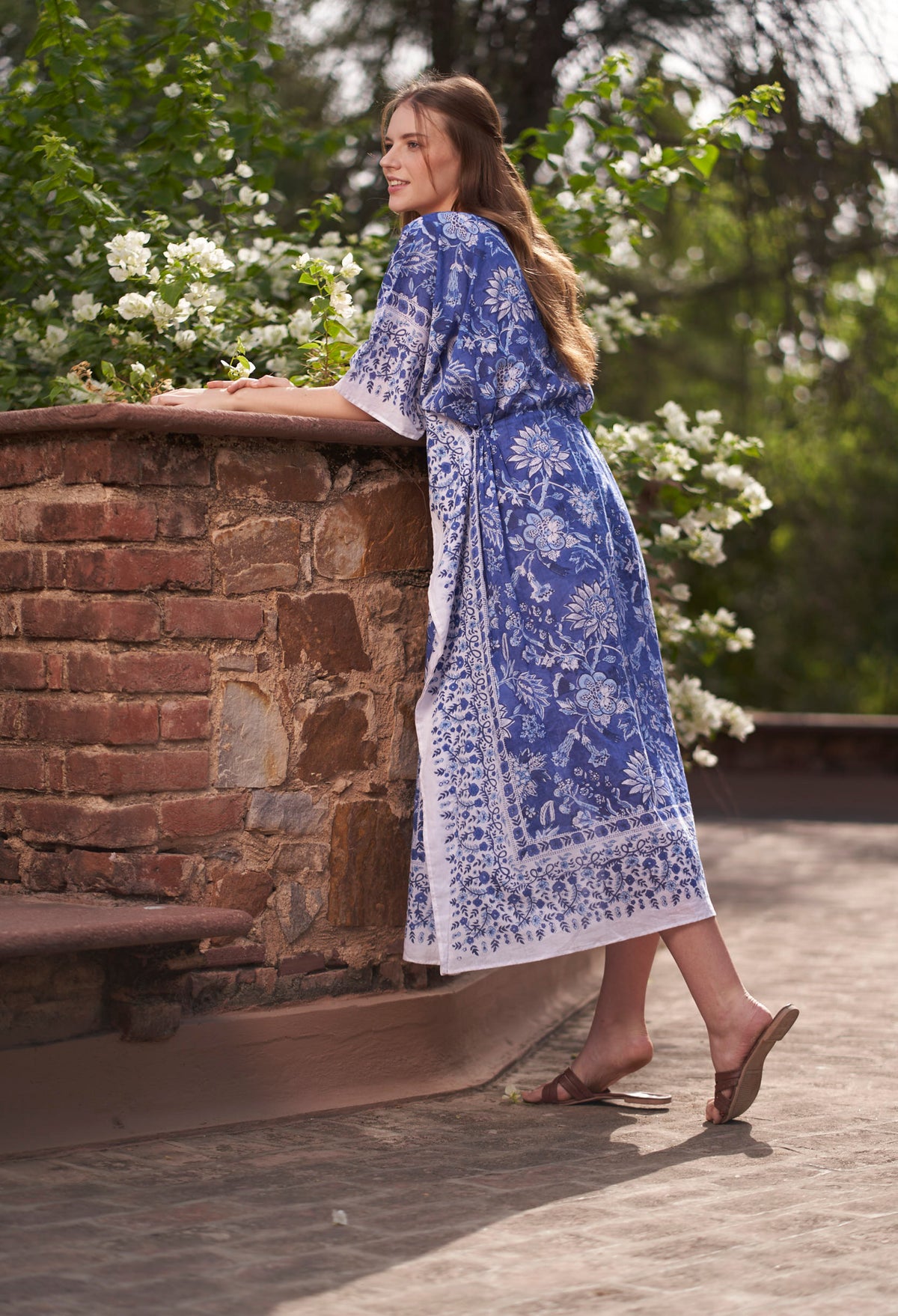 Woman in a blue and white patterned dress standing near a stone wall with greenery in the background