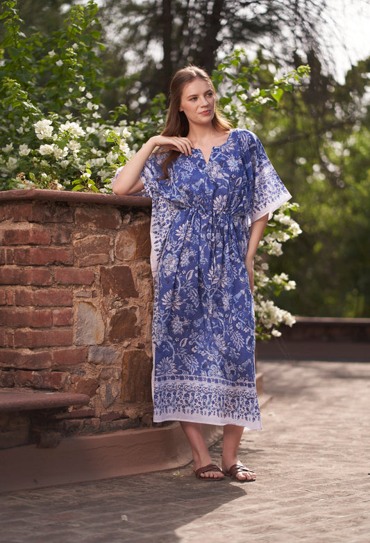 Woman wearing a blue floral dress standing outdoors near a stone wall with greenery.