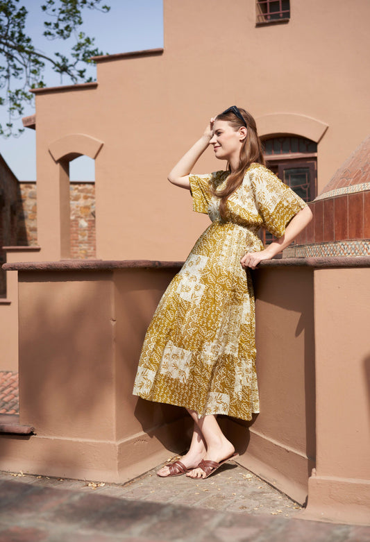 Woman in a mustard green floral dress standing on a balcony with a building in the background