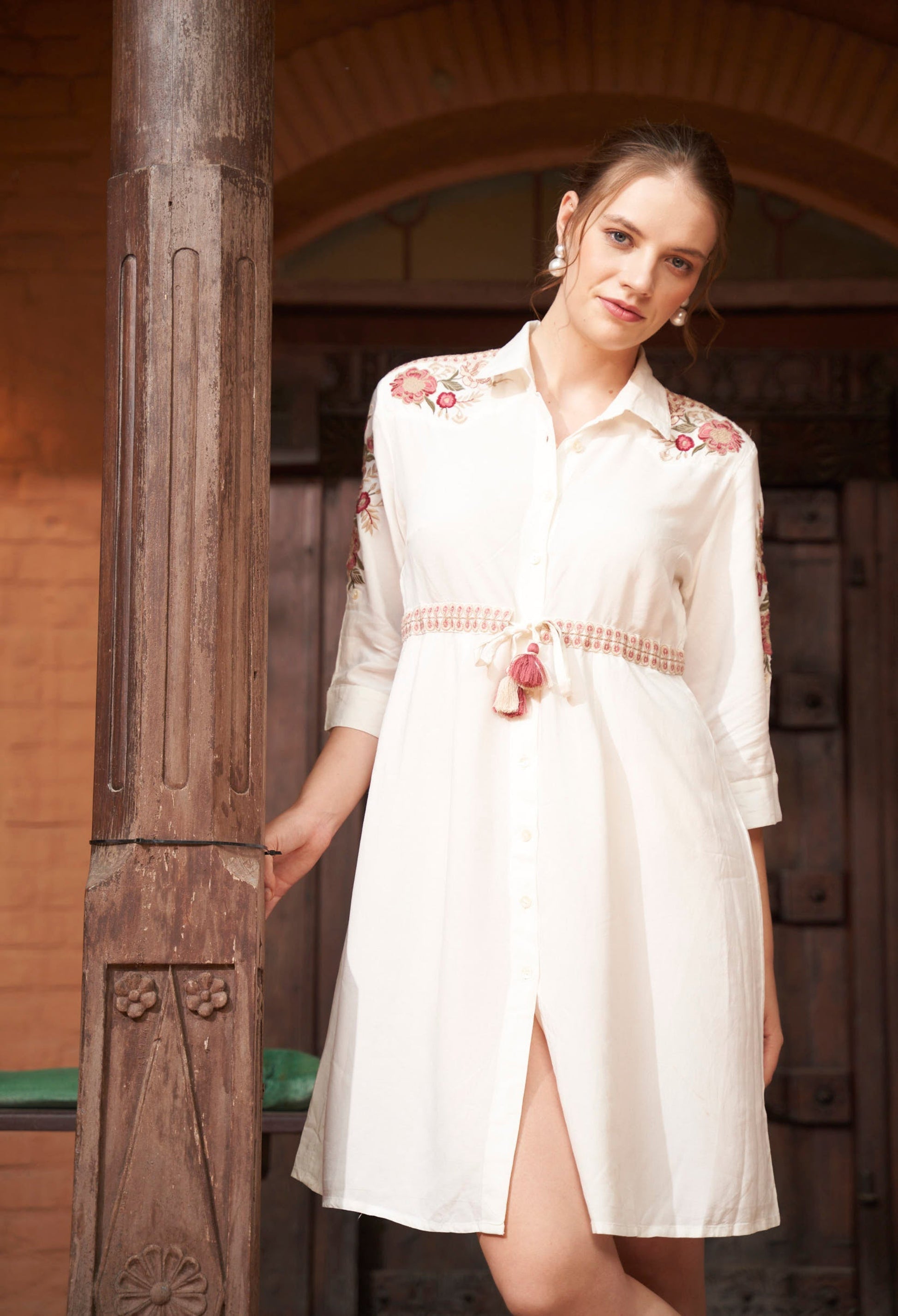 Woman wearing a white dress with floral embroidery standing against a wooden background