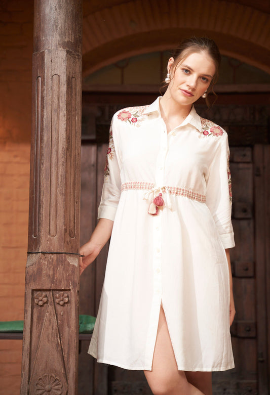 Woman wearing a white dress with floral embroidery standing against a wooden background