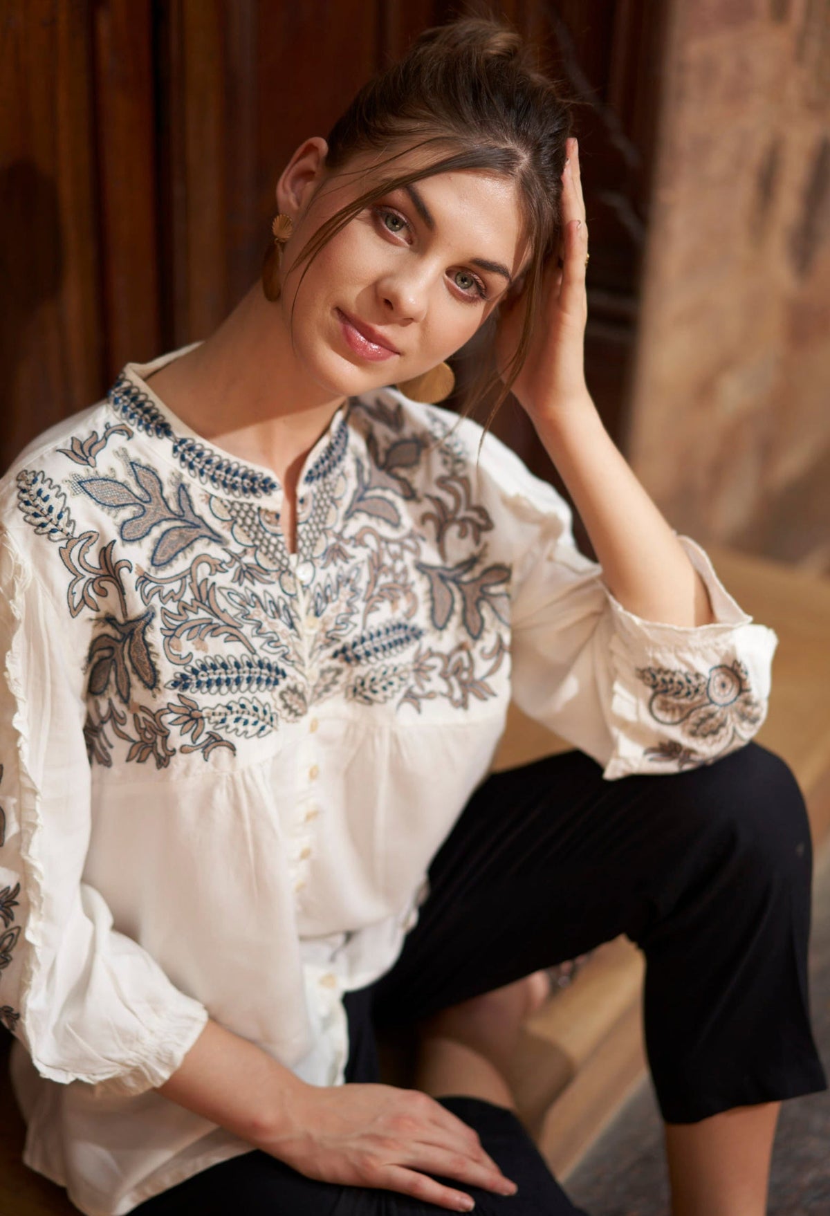 Woman wearing a white blouse with intricate blue embroidery sitting against a wooden wall.