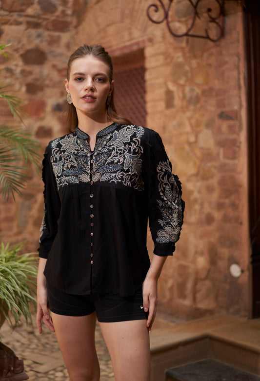 Woman wearing a black blouse with intricate embroidery in front of a stone wall.