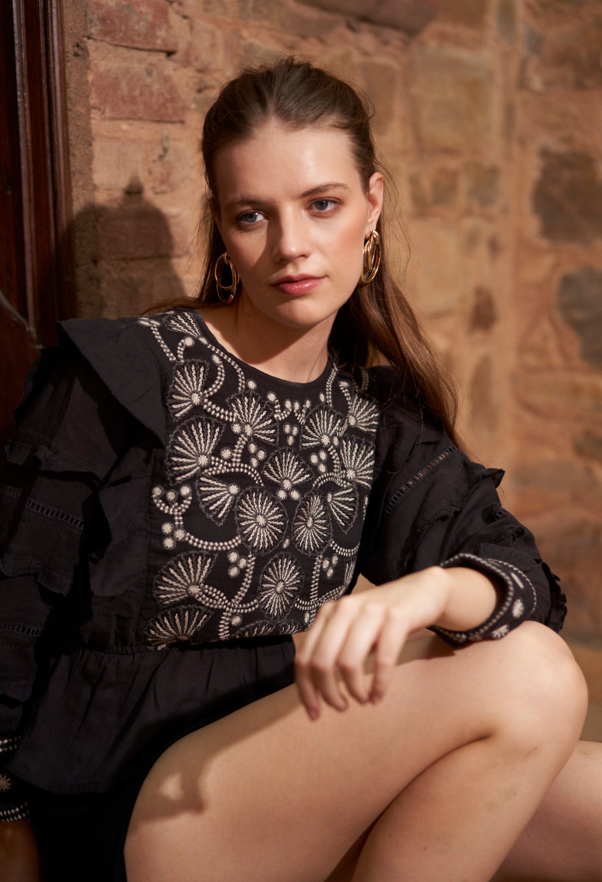 Woman wearing a black top with intricate white patterns against a brick wall.