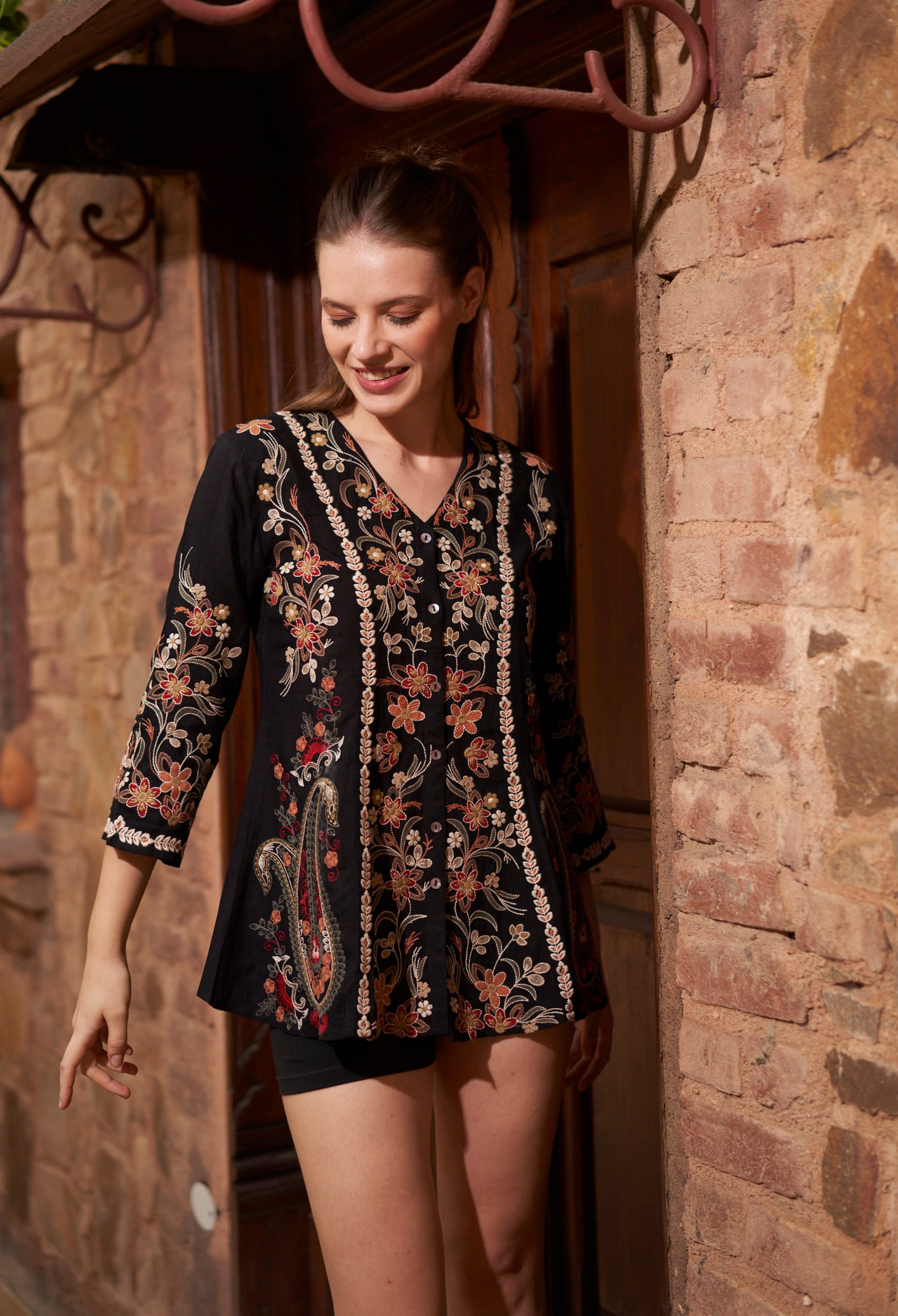 Woman wearing a floral embroidered top standing in front of a stone wall.