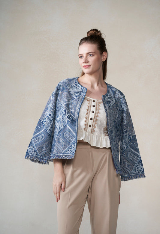 Woman wearing a blue patterned jacket over a white blouse with beige pants against a plain background