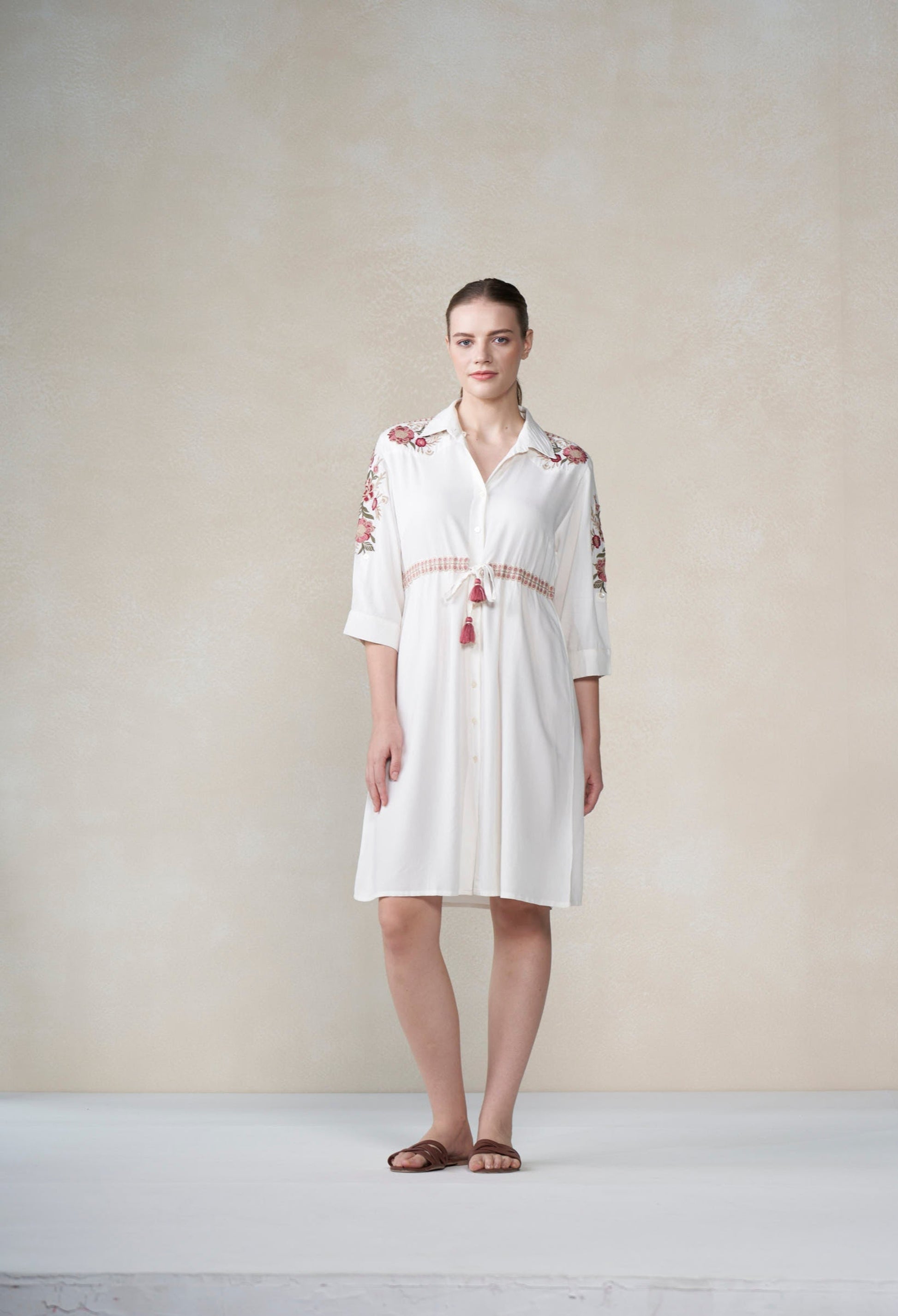 Woman wearing a white dress with red embroidery against a plain background