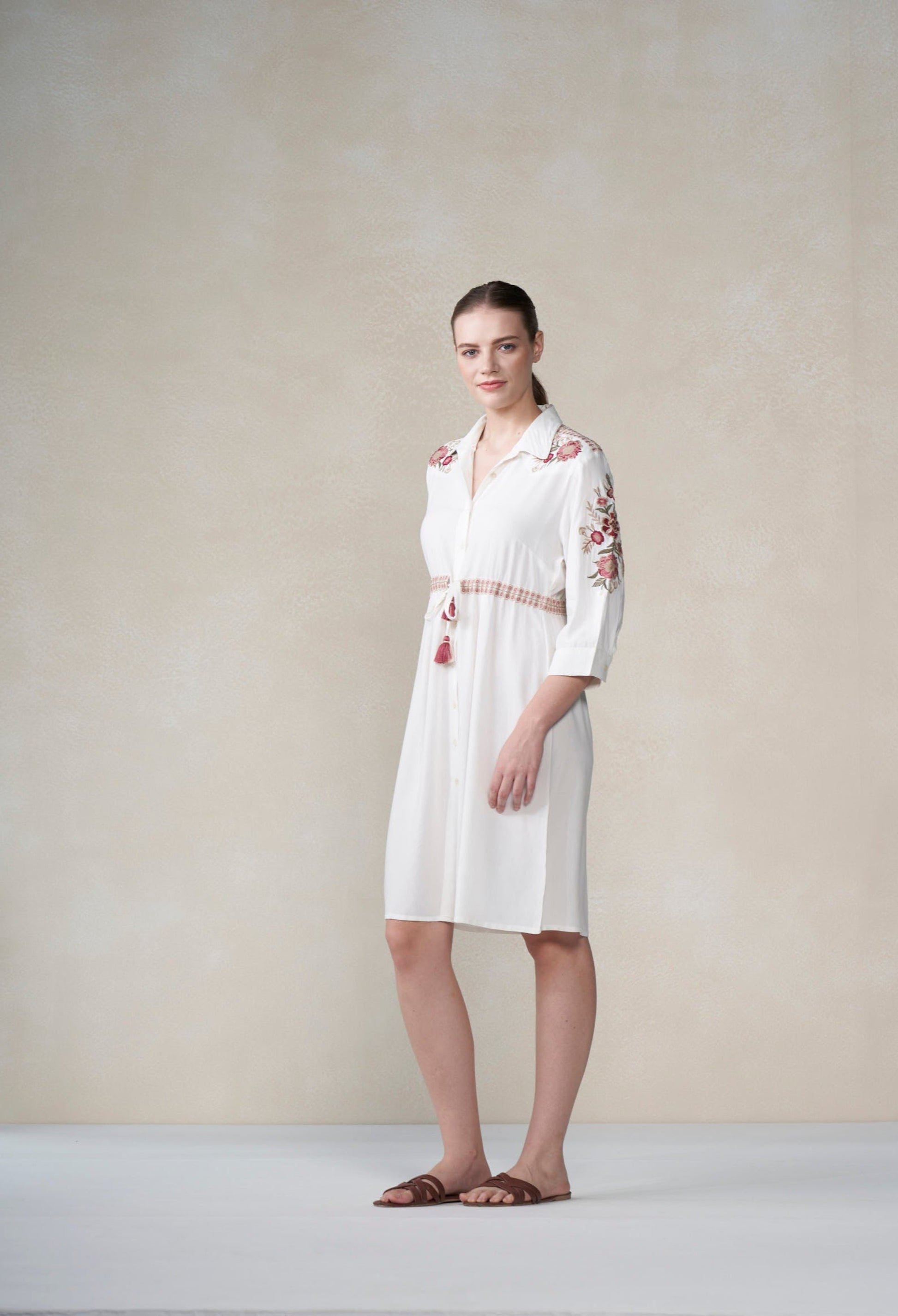 Woman wearing a white dress with floral embroidery against a plain background