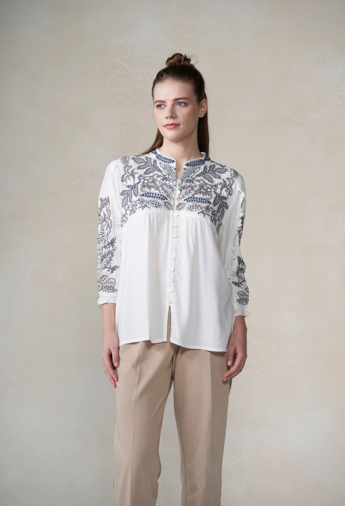 Woman wearing a white blouse with blue floral embroidery and beige pants against a plain background
