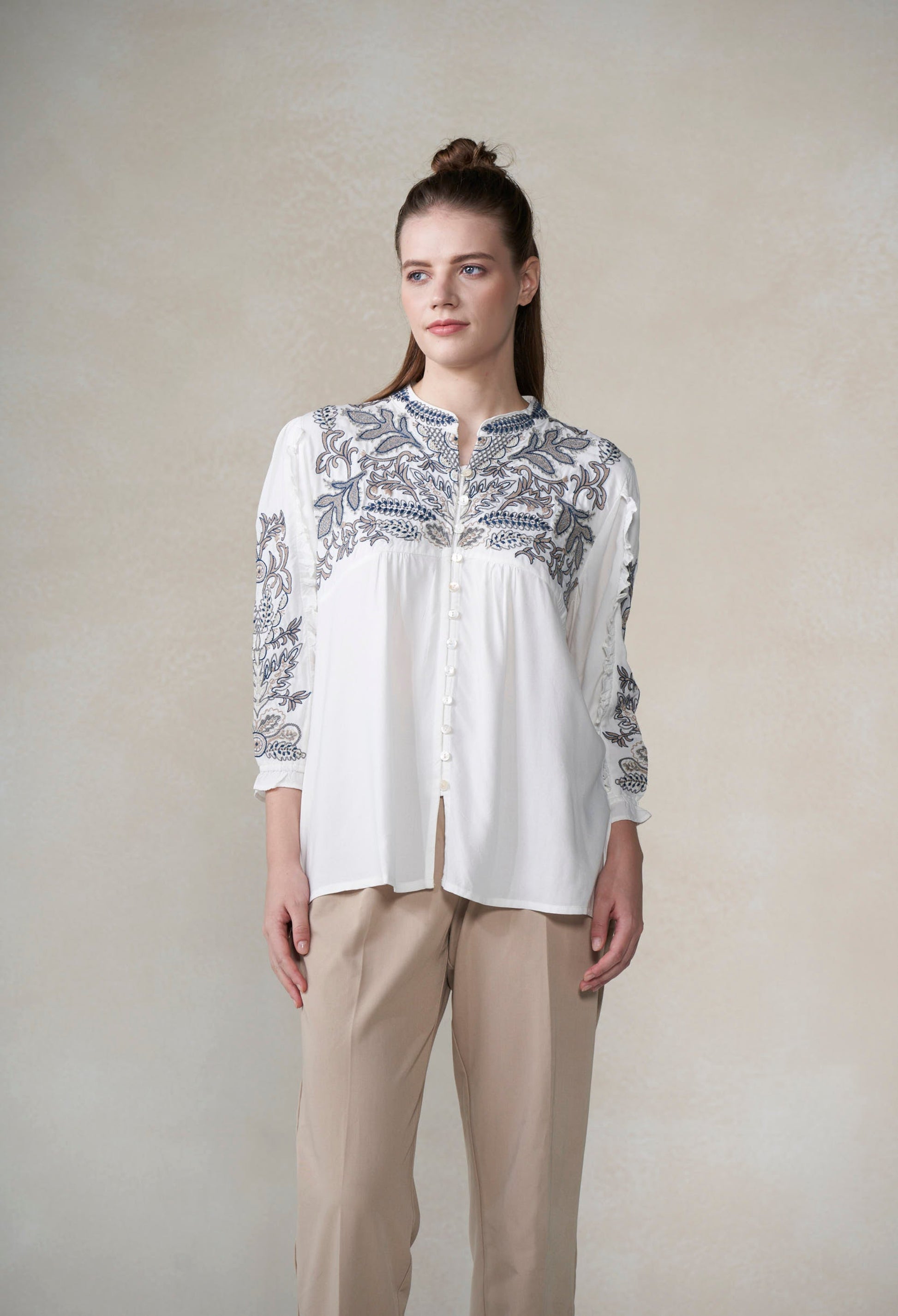 Woman wearing a white blouse with blue floral embroidery and beige pants against a plain background