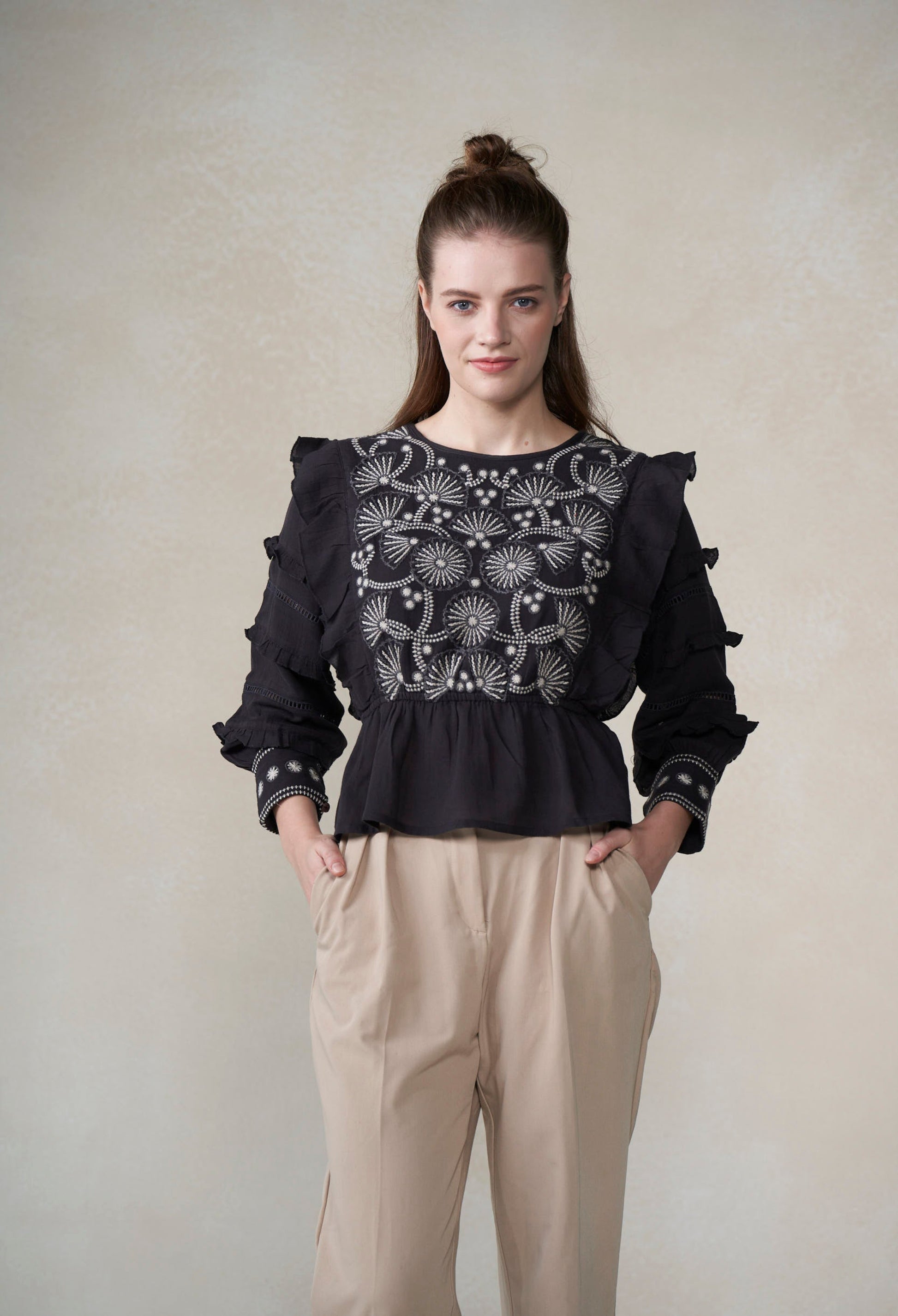 Woman wearing a black embroidered blouse and beige pants against a plain background