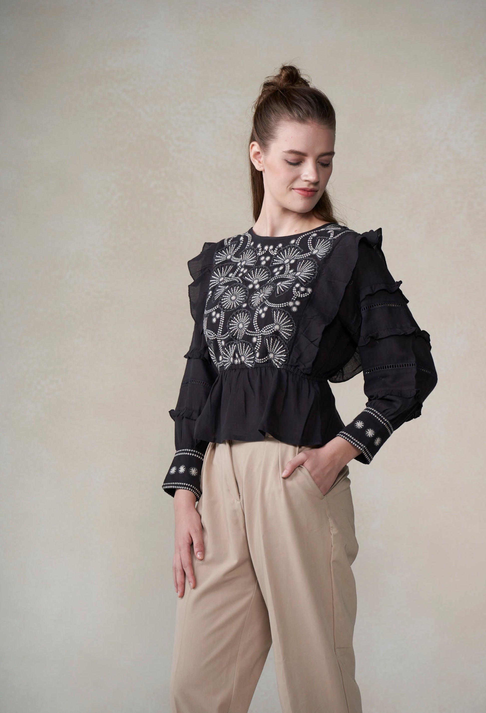 Woman wearing a black embroidered blouse and beige pants against a plain background