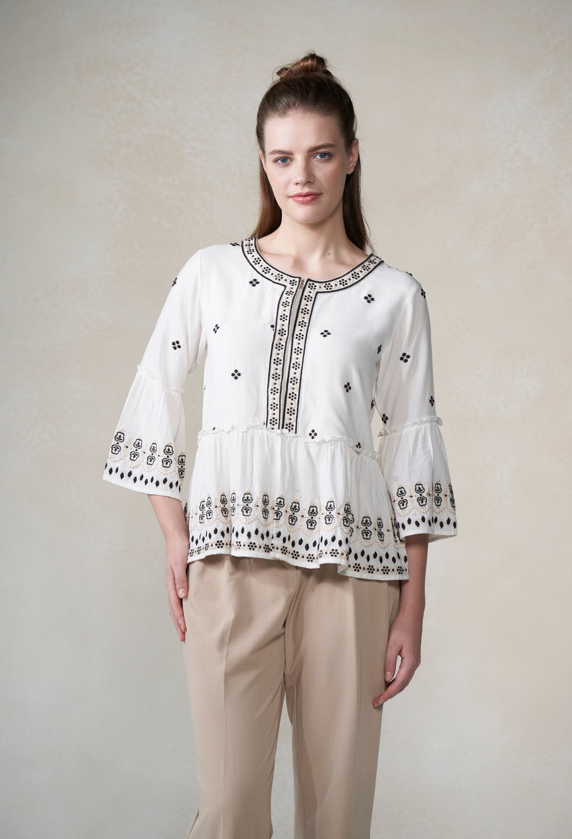 Woman wearing a white blouse with black embroidery and beige pants against a plain background