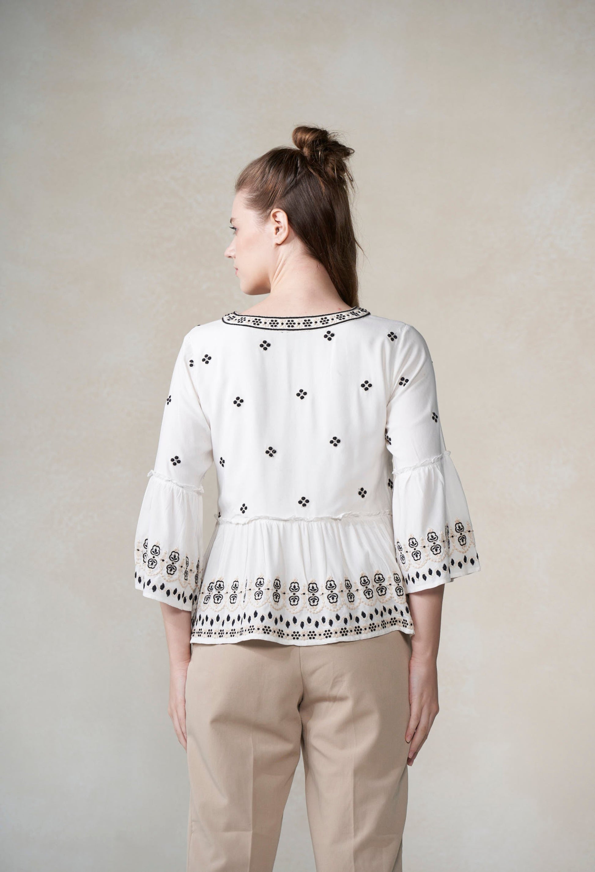 Woman wearing a white blouse with black patterns and beige pants against a plain background