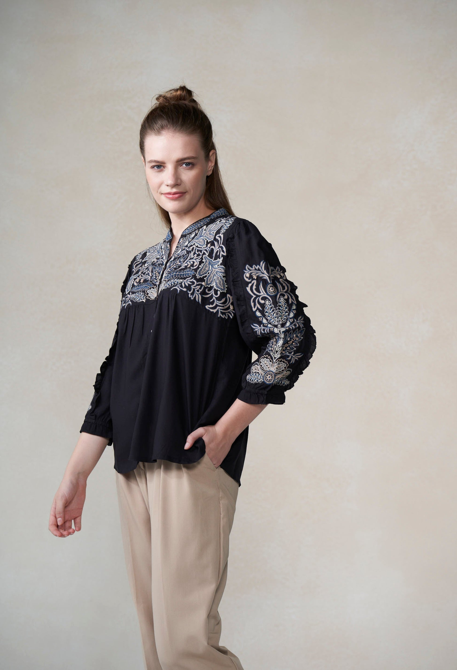 Woman wearing a black blouse with white  and blue embroidery on a plain background