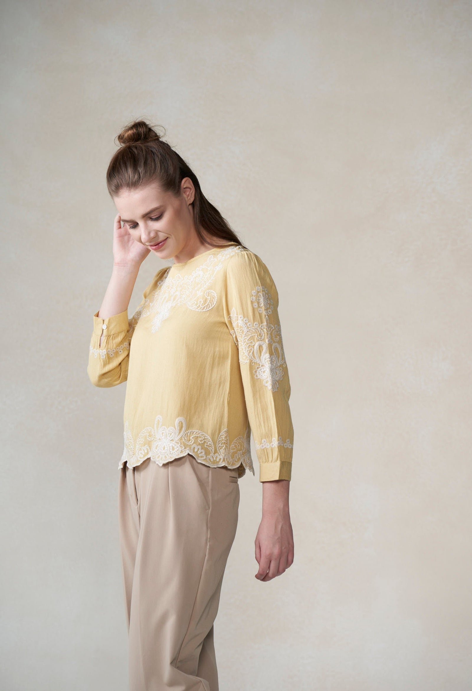 Woman wearing a light yellow blouse with lace details and beige pants against a plain background