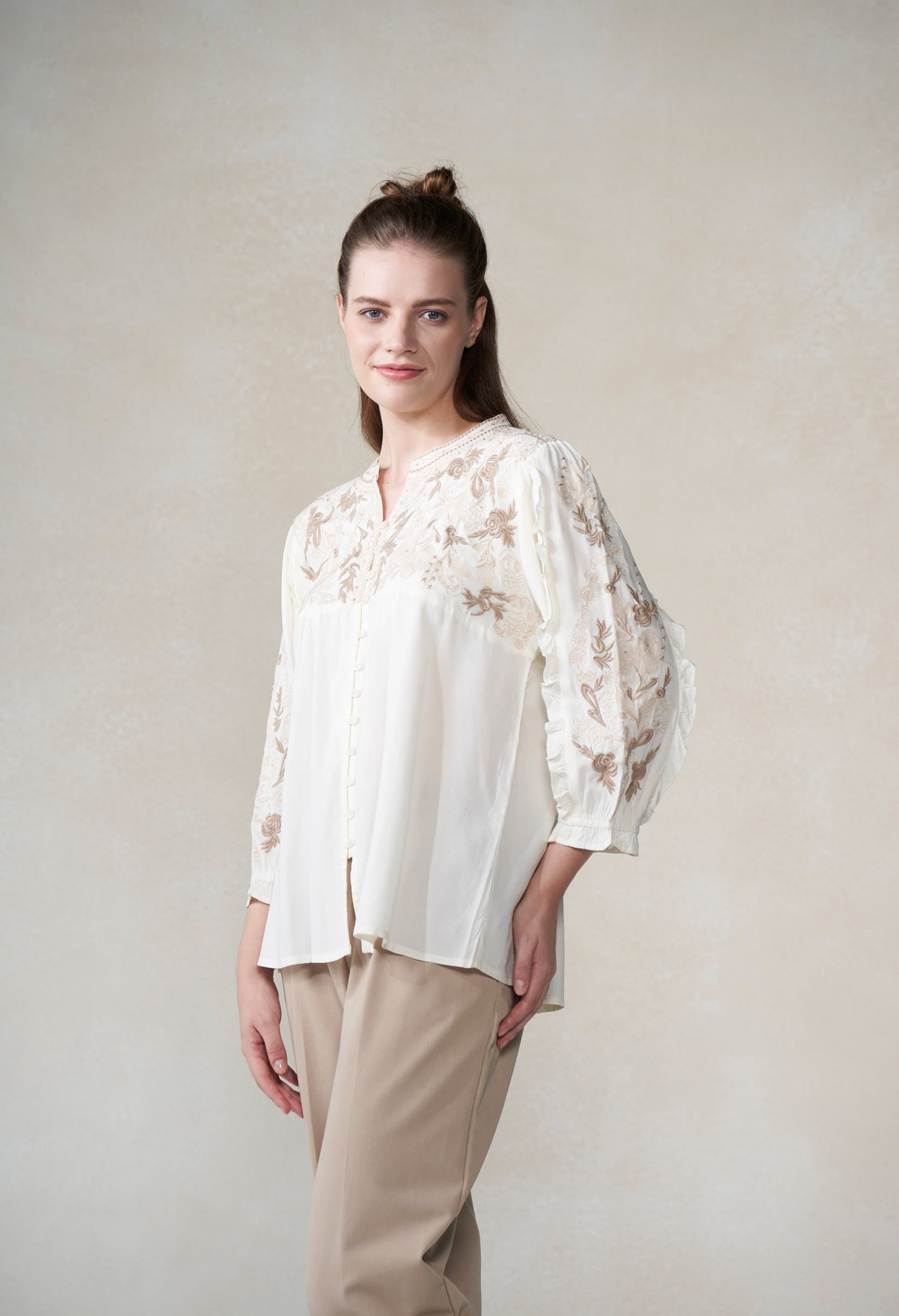 Woman wearing a white blouse with floral patterns and beige pants against a plain background