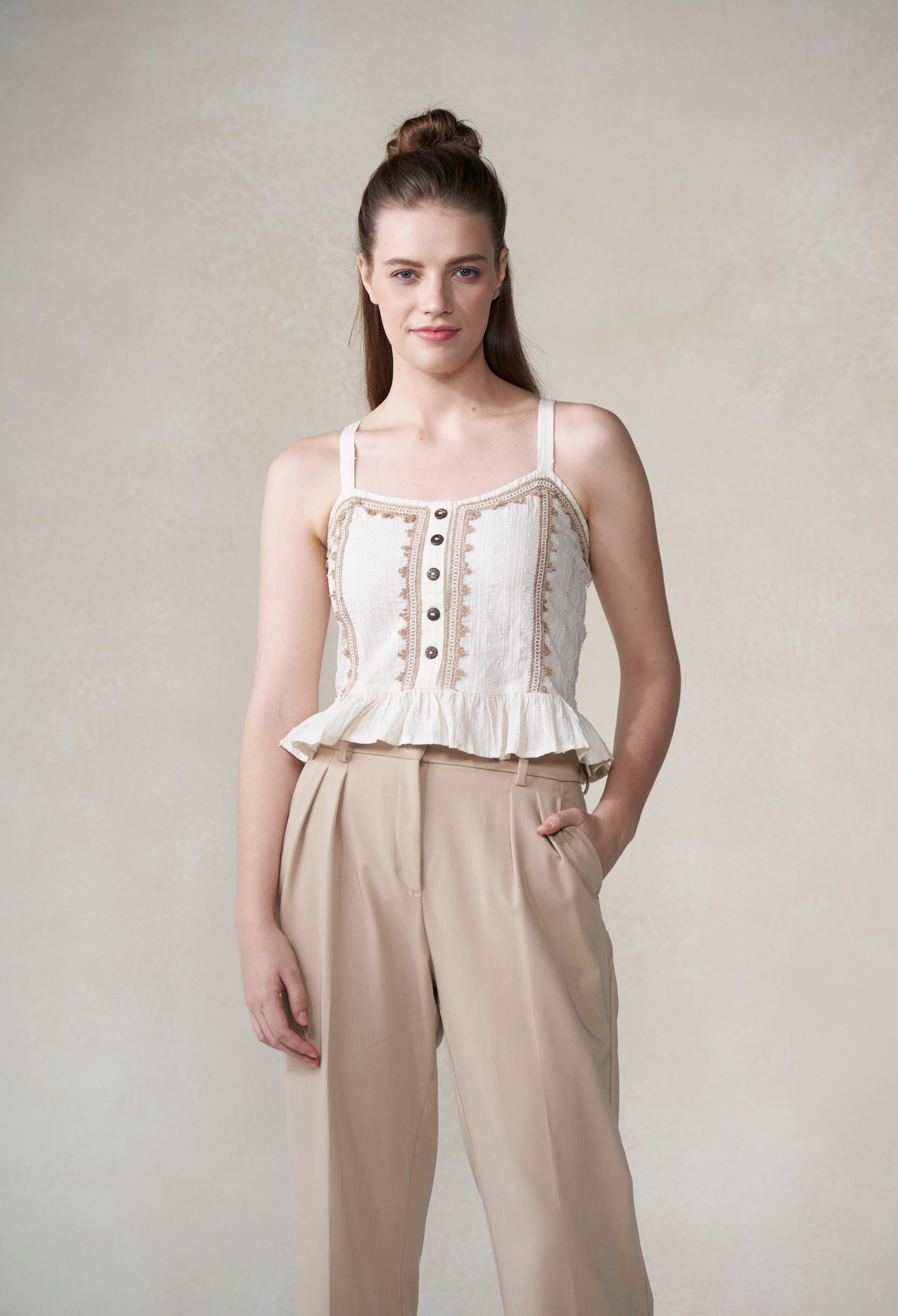Woman wearing a white top with beige lace details and beige pants against a plain background