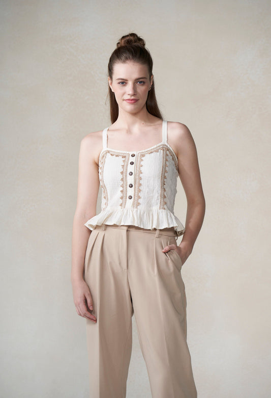 Woman wearing a white top with beige lace details and beige pants against a plain background