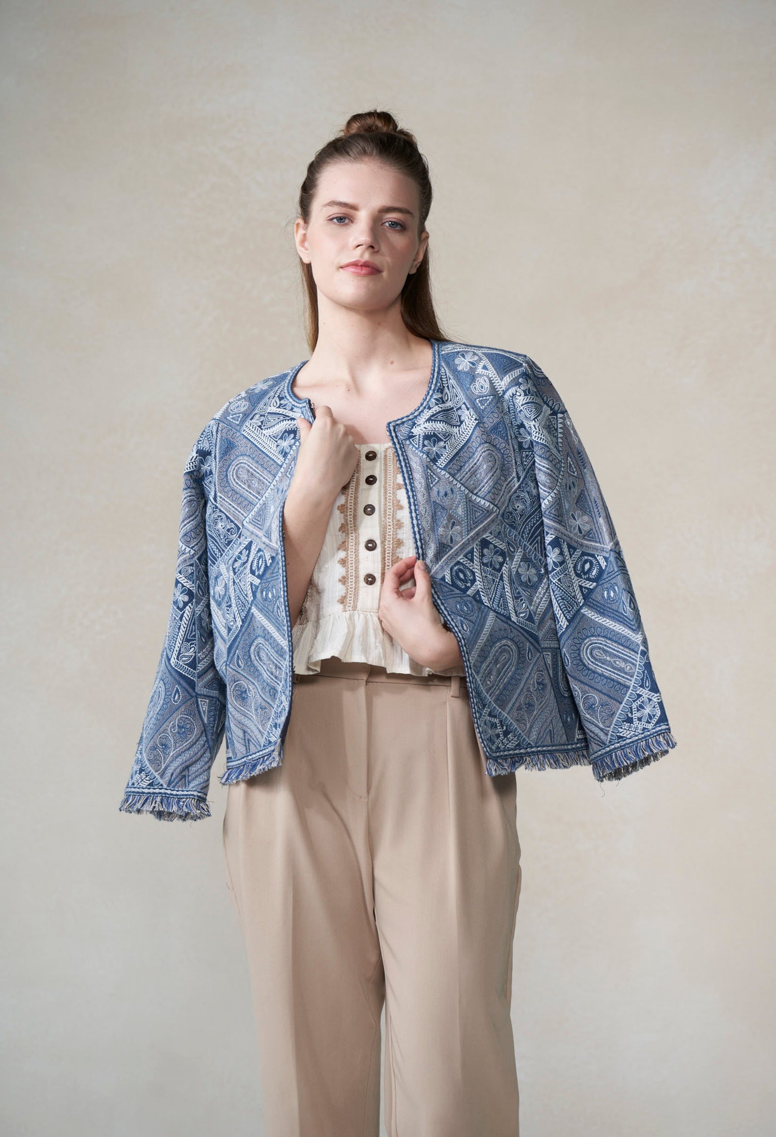 Woman wearing a blue patterned jacket over a white top and beige pants on a plain background