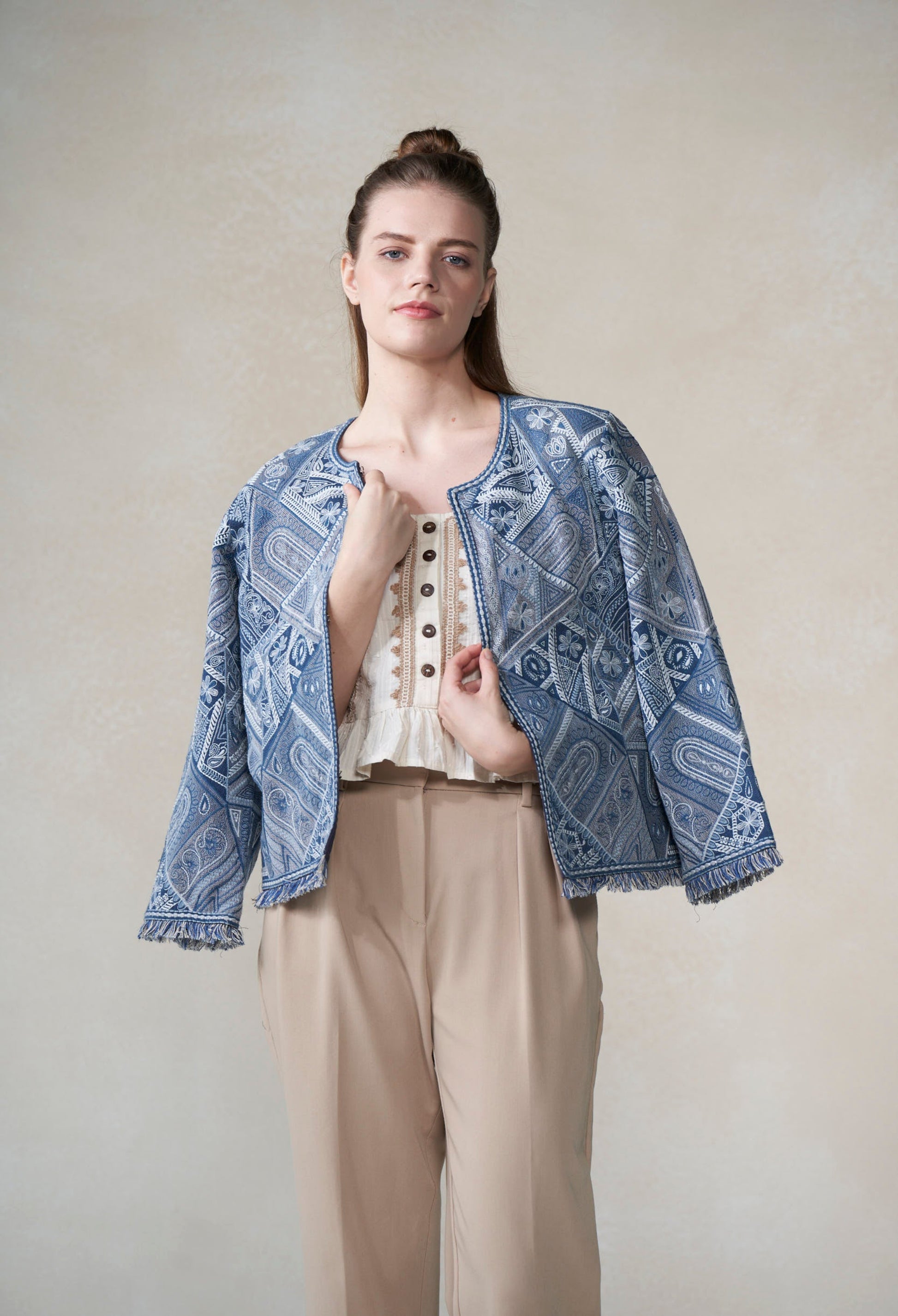 Woman wearing a blue patterned jacket over a white top and beige pants on a plain background