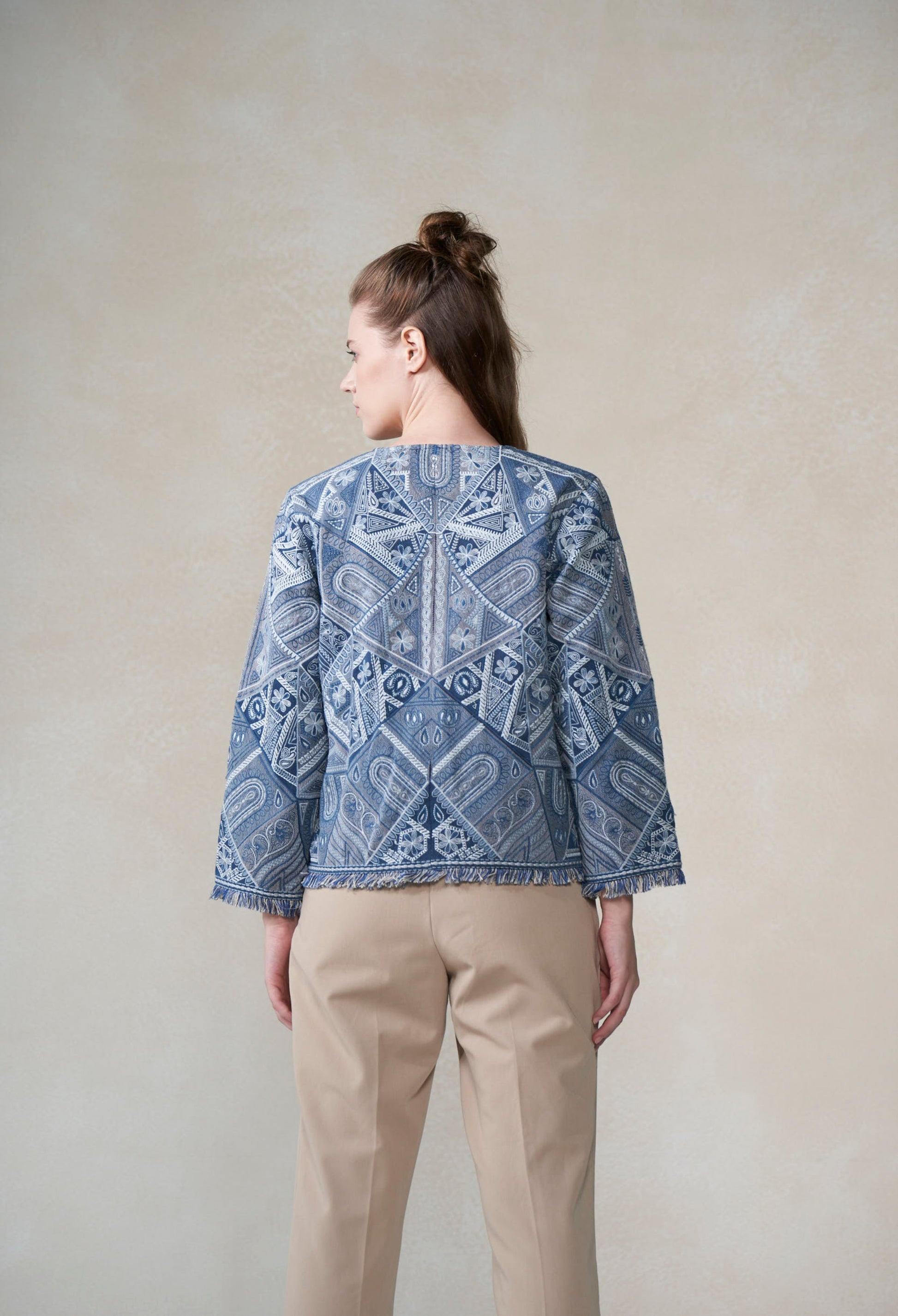 Woman wearing a blue patterned jacket with white embroidery and beige pants against a plain background