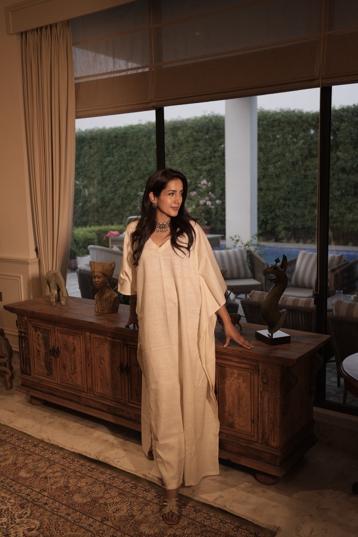 A girl wearing a ivory khadi silk kaftan by my marigold summer , she is standing in a living room in delhi. 