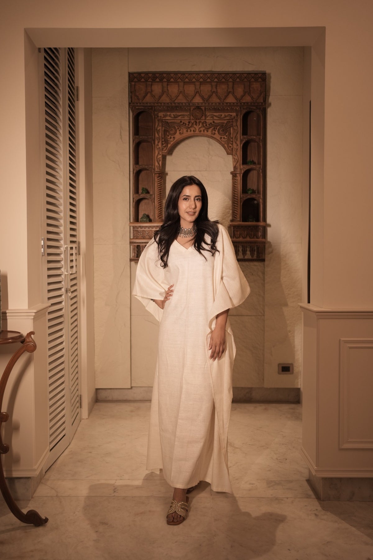 a girl wearing a ivory khadi silk kaftan by my marigold summer brand , she is standing in front of an ancient looking window in a house in delhi. 
