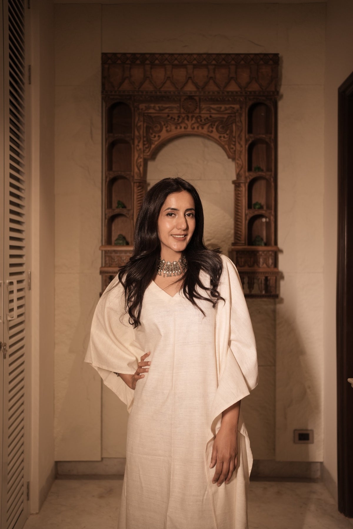 a girl wearing a ivory khadi silk kaftan by my marigold summer brand , she is standing in front of an ancient looking window in a house in delhi. 