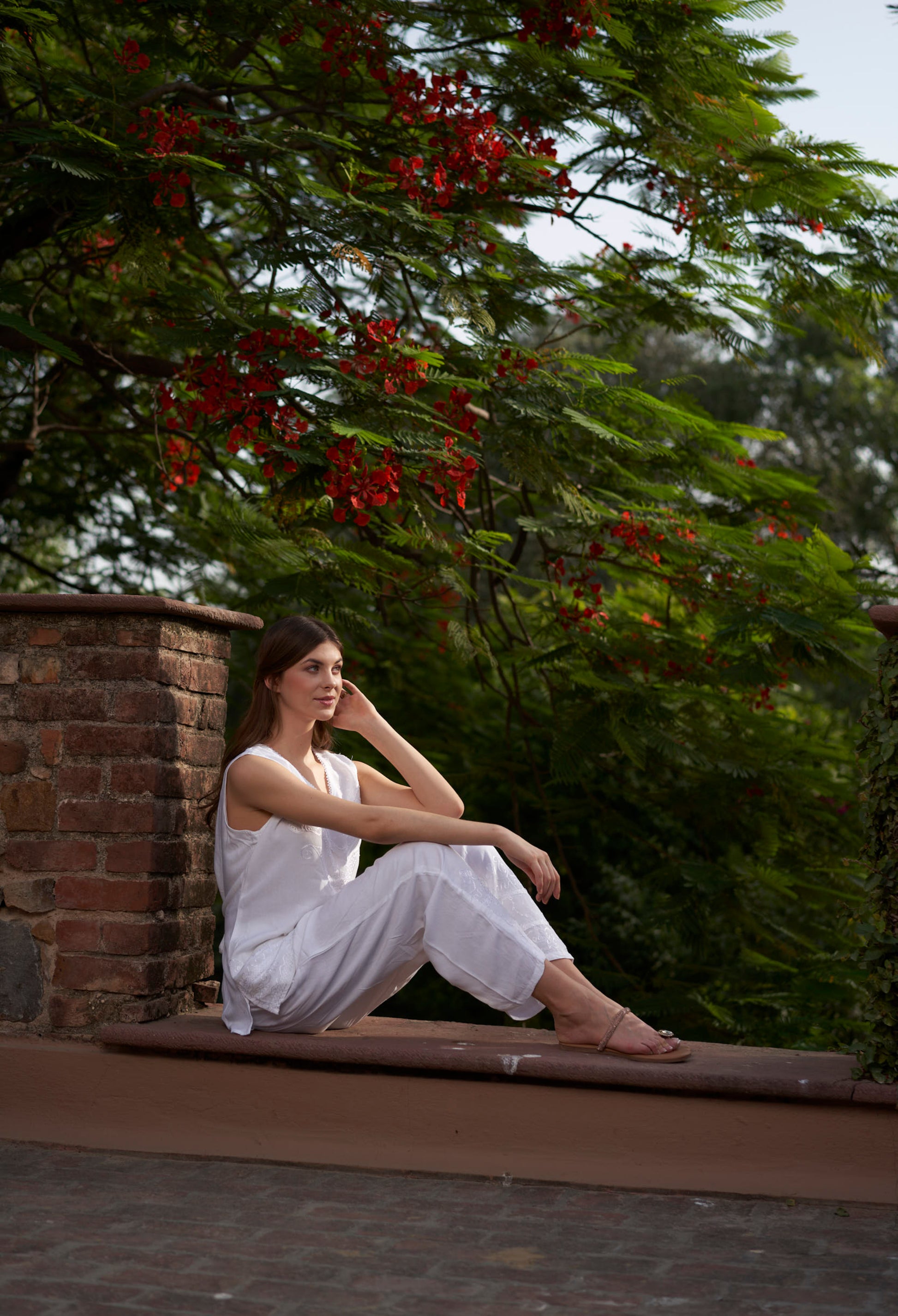 Woman in a white outfit sitting on a stone ledge with greenery and red flowers in the background