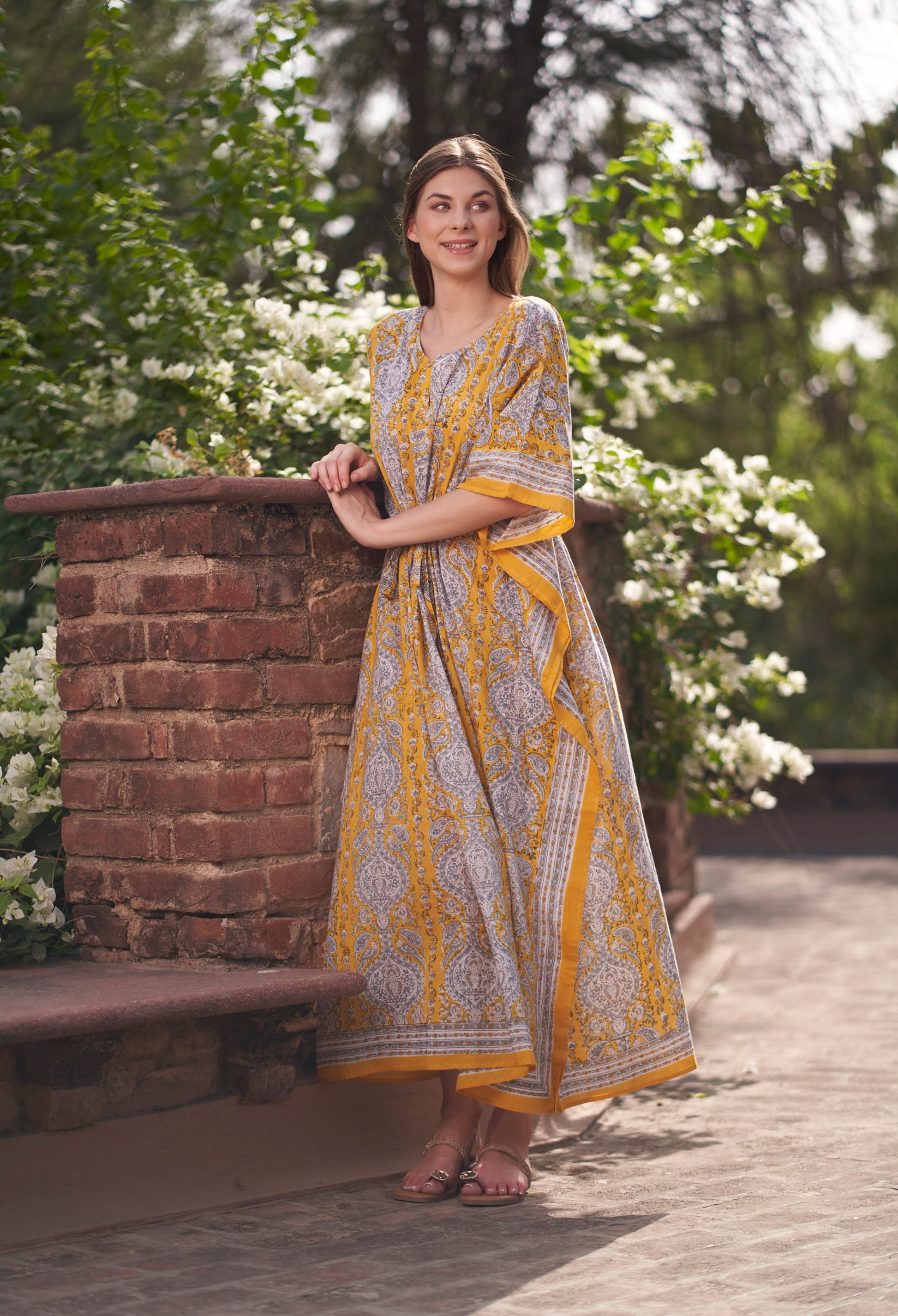 Woman in a yellow and white patterned dress standing outdoors with greenery in the background