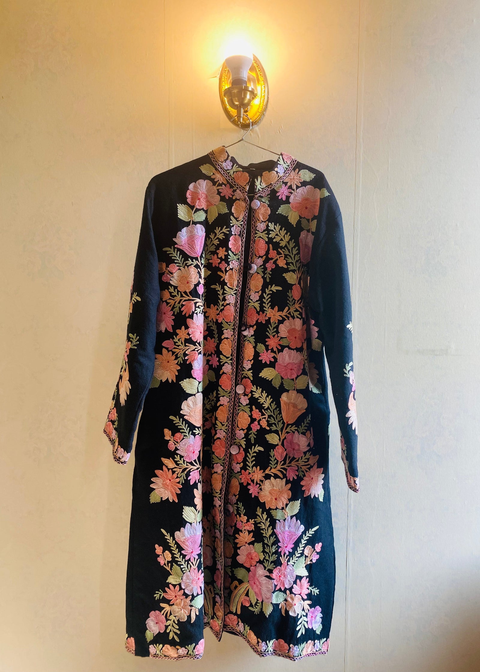 Embroidered Kashmiri phiran in black with pink and gold floral motifs, displayed under a wall light. Front image. 
